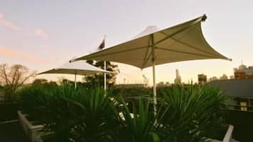 A rooftop terrace with large umbrellas providing shade, surrounded by green plants.