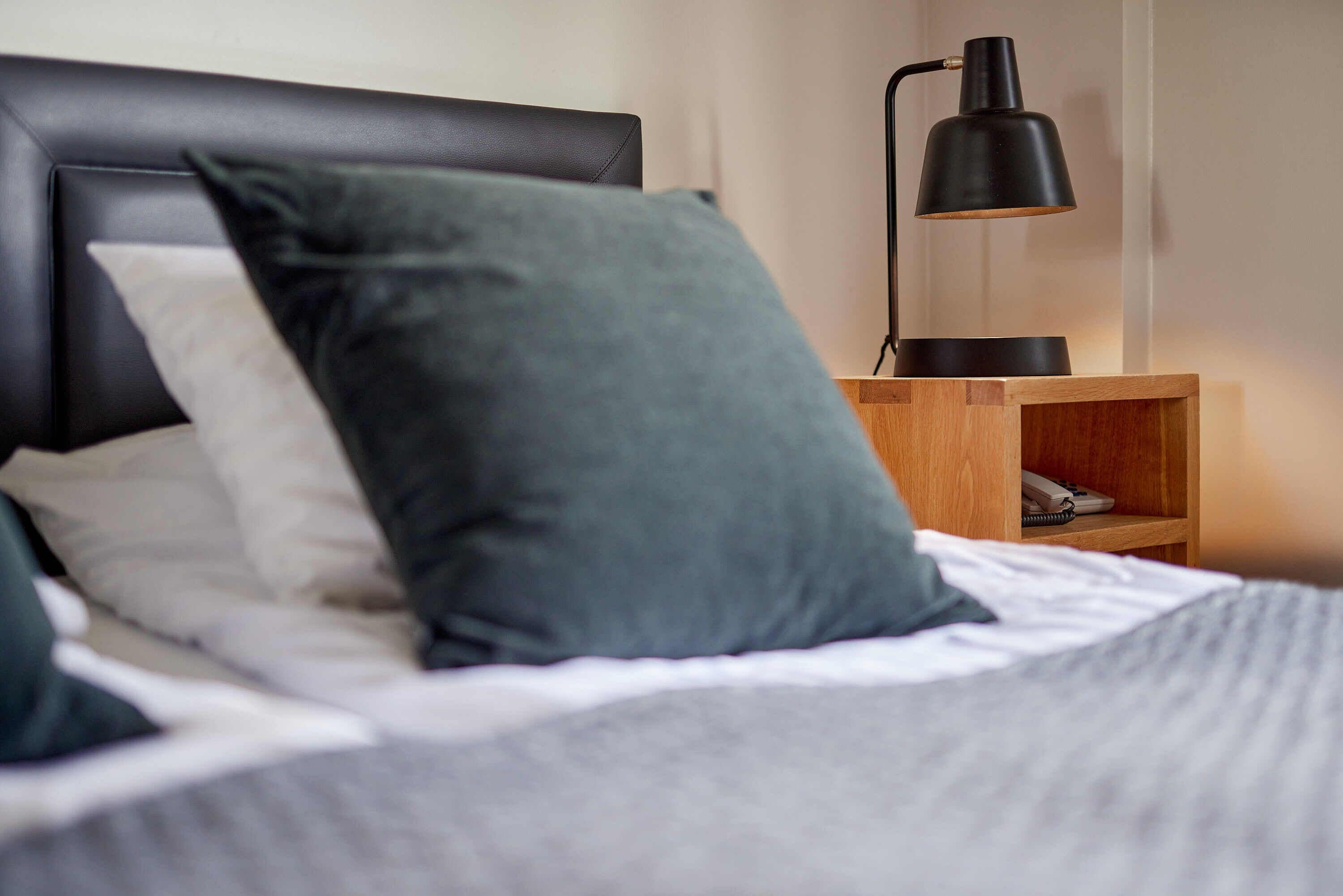 A neatly made bed with a black headboard, a bedside table with a lamp, and a wooden shelf with a phone and a book.