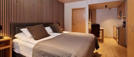Un dormitorio moderno con una cama grande, paneles de madera en la pared, un escritorio pequeño y una kitchenette al fondo.