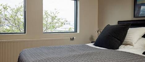 Un dormitorio con una cama, dos ventanas, un radiador y un cuadro en la pared.