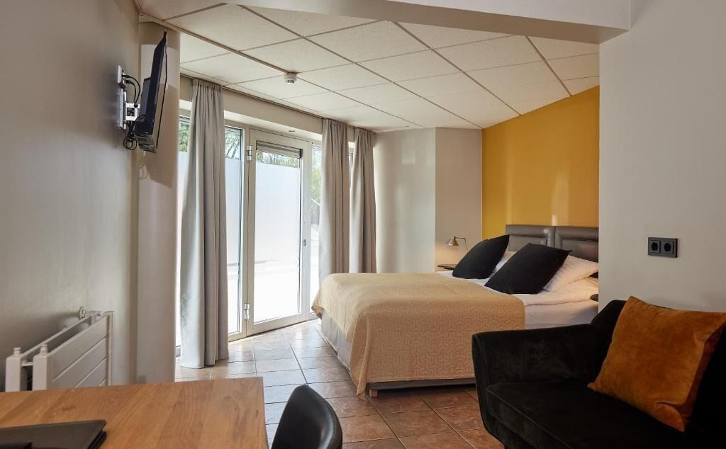 Habitación de hotel con cama, sofá, televisor montado en la pared y ventana con cortinas.