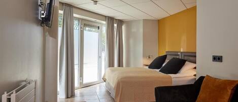 Habitación de hotel con cama, sofá, televisor montado en la pared y ventana con cortinas.