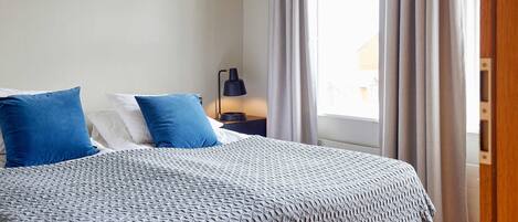 Una cama bien hecha con una colcha estampada, dos almohadas y una mesita de noche con una lámpara.