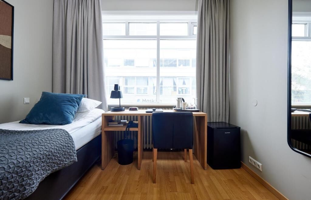A hotel room with a bed, a desk, a chair, a radiator, and a window with curtains.