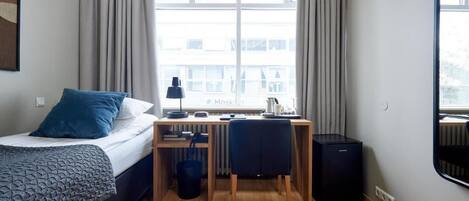 A hotel room with a bed, a desk, a chair, a radiator, and a window with curtains.