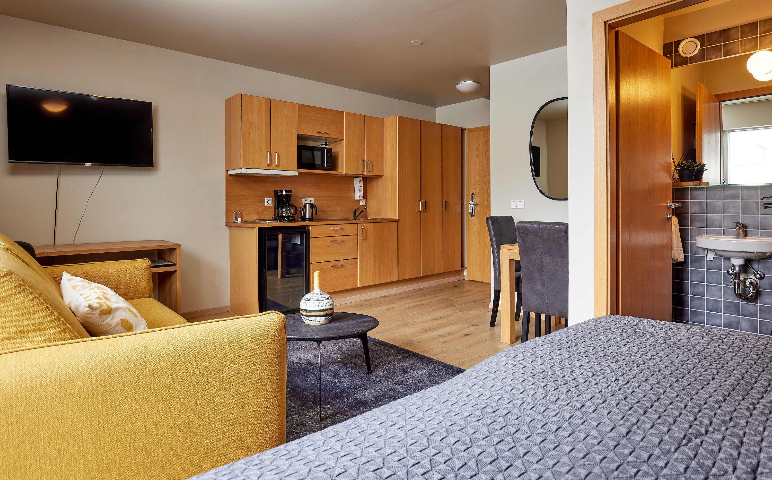Una habitación de hotel moderna con sofá, una mesita, cama, zona de cocina con armarios de madera, televisor de pantalla plana y baño con lavabo y espejo.