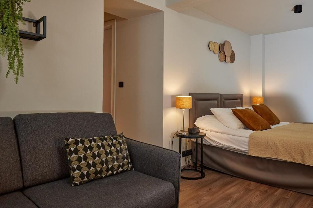 A modern bedroom with a grey sofa, a bed with a beige blanket, two lamps, and a wall-mounted decorative piece.