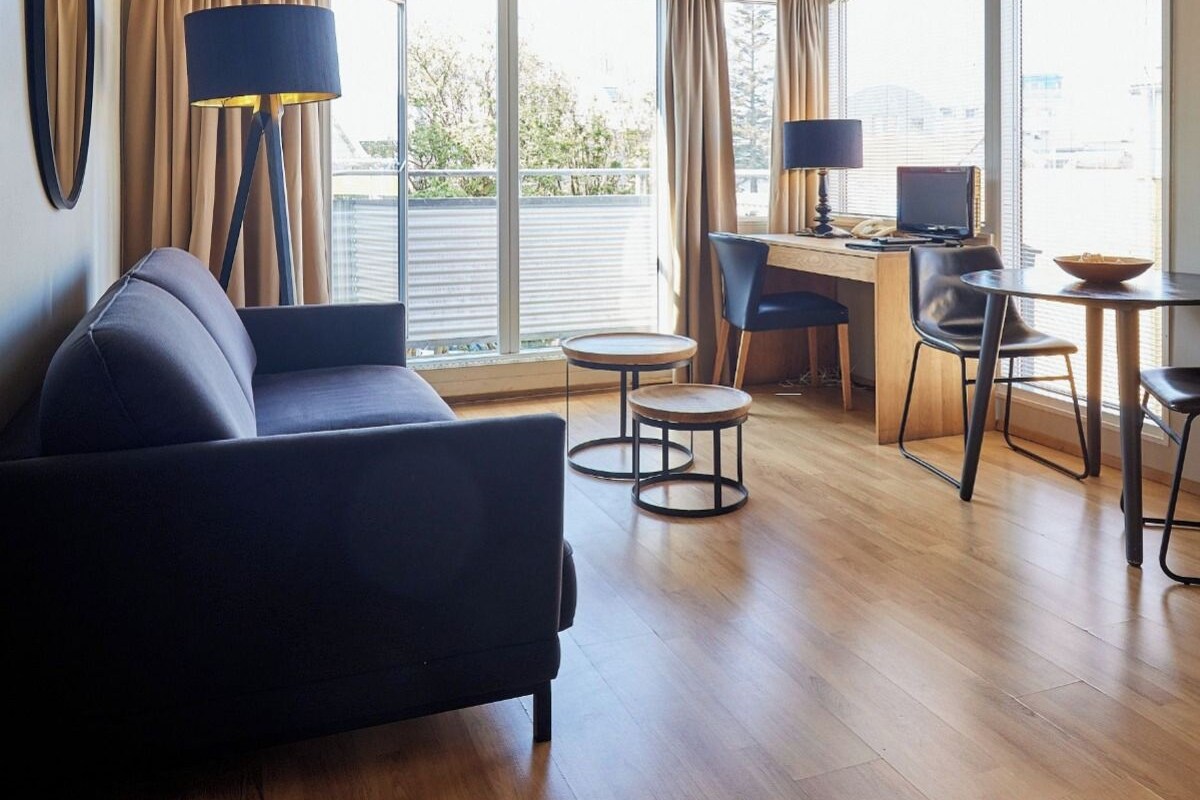 A modern living room with a sofa, two small round tables, a desk with a computer, and large windows with curtains.