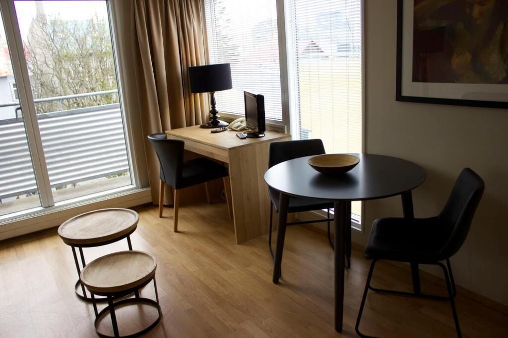 A room with a desk, two chairs, a small table, and a window with curtains.