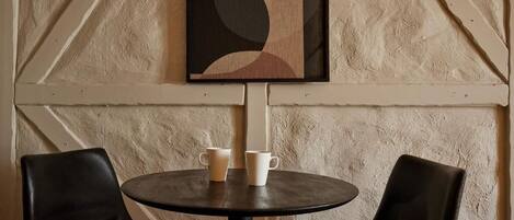 Une table ronde avec deux chaises noires, une œuvre d’art abstraite encadrée au-dessus, et deux tasses blanches posées sur la table.