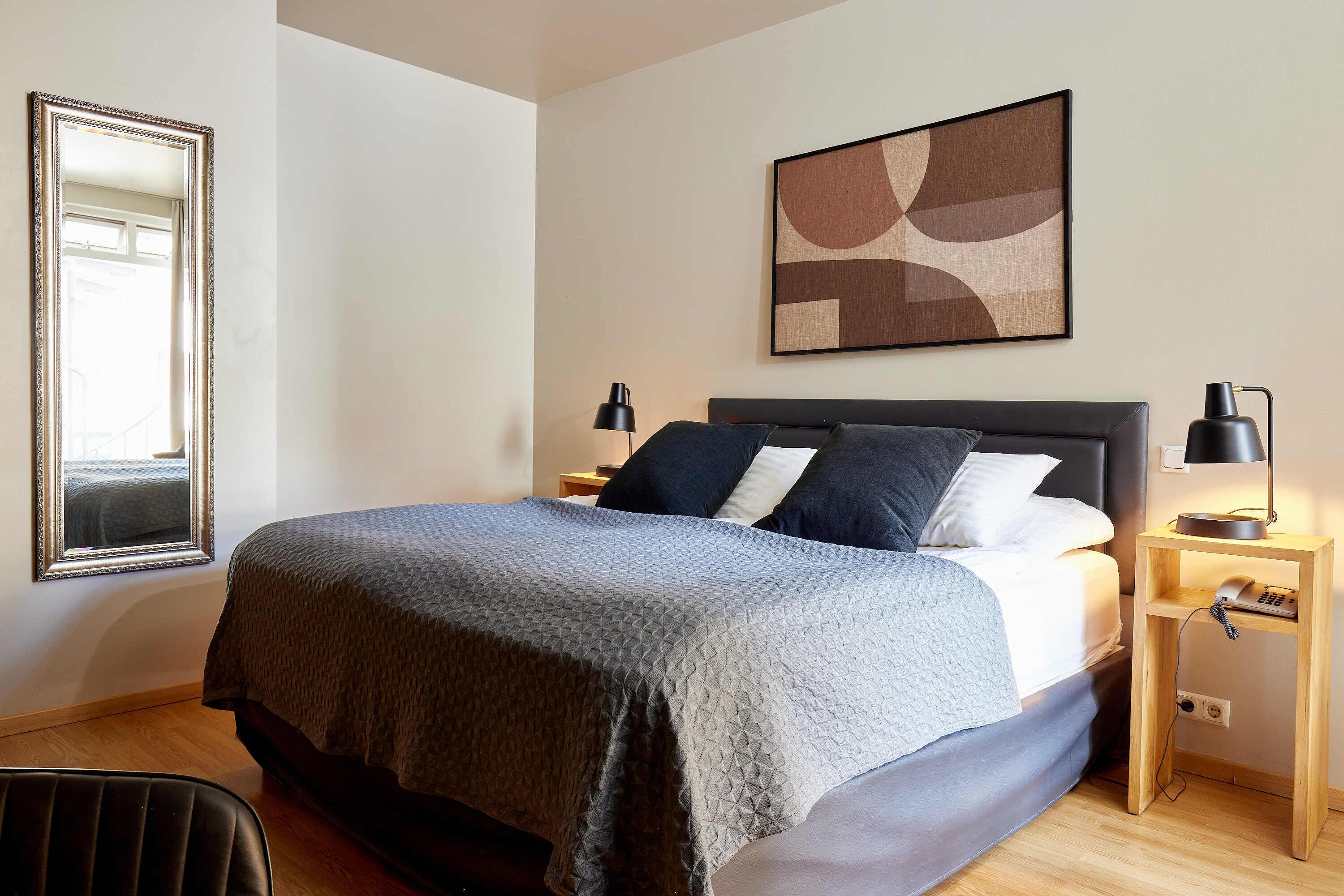 A neatly made bed with a grey quilt, two bedside lamps, a wooden nightstand, and a framed abstract painting on the wall.