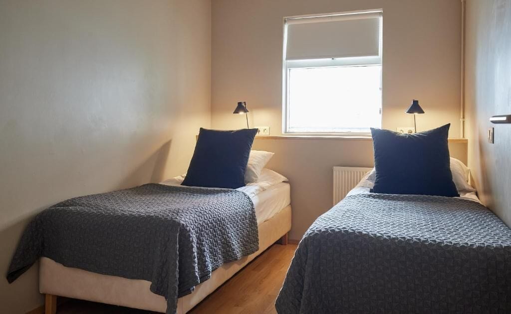 A room with two beds, each with a blue pillow and a grey blanket. There is a window with blinds, a radiator, and a lamp on each bed.