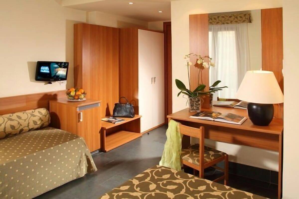 A hotel room with a bed, a desk with a lamp, a chair, a TV, and a wardrobe.