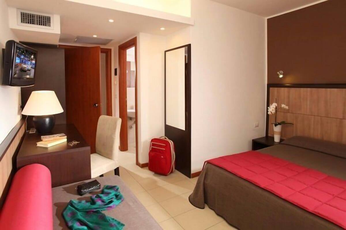 A hotel room with a bed, a desk with a lamp, a chair, a TV, and a red suitcase.