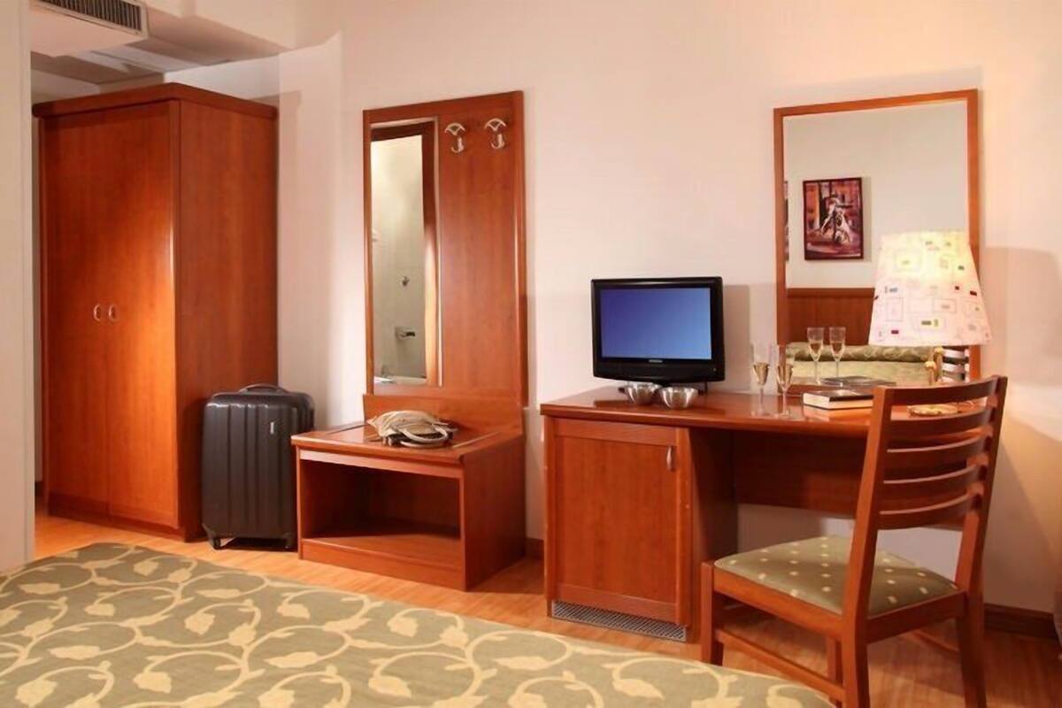 A hotel room with a wooden wardrobe, a desk with a television, a chair, a small table with a lamp, and a bed with a patterned bedspread.