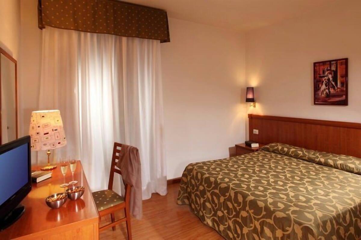 A hotel room with a wooden desk, a television, a bed with patterned bedding, a chair, a bedside table, and a wall-mounted lamp.