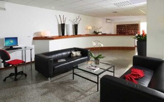 Un moderno vestíbulo con un sofá de cuero negro, una mesa de centro de cristal, un escritorio con computadora y una recepción con mostrador de madera y plantas decorativas.