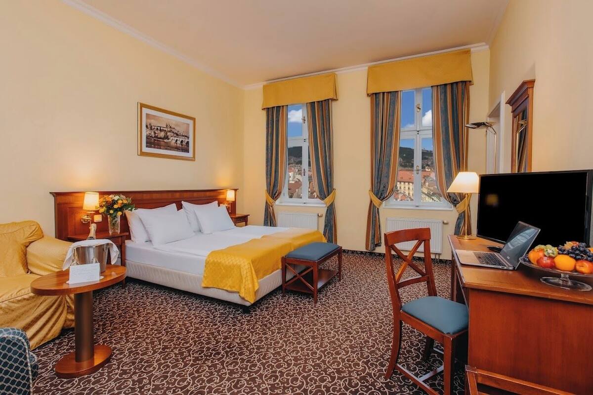 A hotel room with a bed, a desk with a laptop, a chair, a small table with a lamp, and a view of the city through the windows.