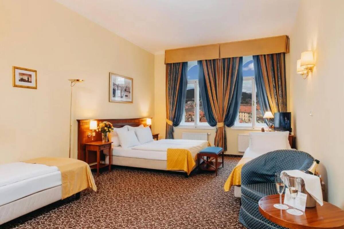 A hotel room with a bed, a sofa, a small table with glasses, and a window with curtains.