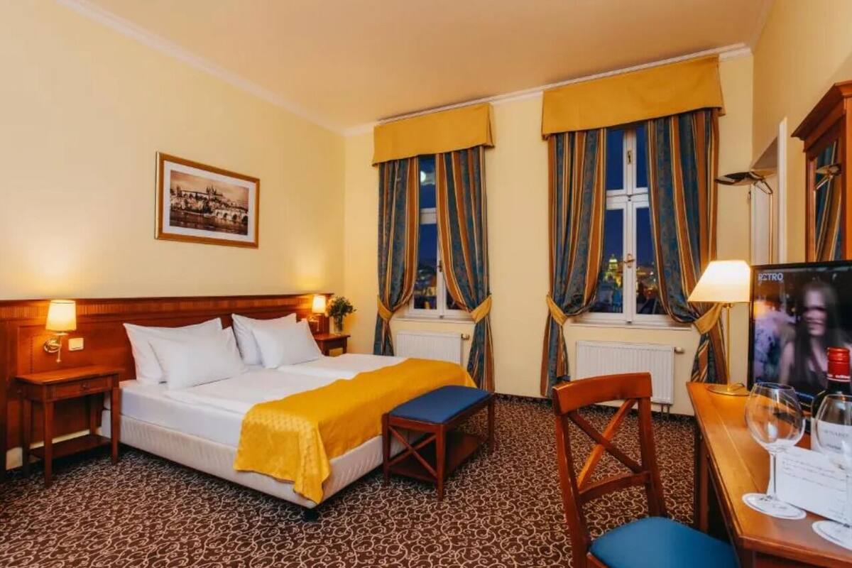 A hotel room with a bed, a desk with a chair, a window with curtains, a picture on the wall, and a television.