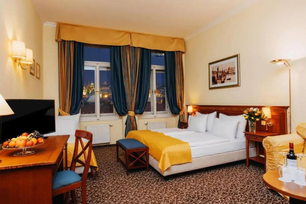 A hotel room with a bed, a desk with a fruit bowl, a chair, a table with a bottle and glasses, a TV, and a window with curtains.