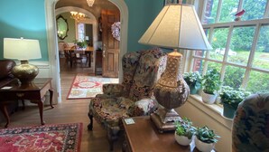 Un salon avec un fauteuil à motifs floraux, une table basse en bois et un tapis rouge à motifs. Une fenêtre offre une vue sur la verdure à l’extérieur.