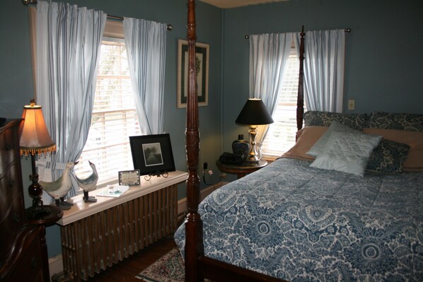 Un dormitorio con cama con dosel, mesita de noche con lámpara, radiador y ventana con cortinas.
