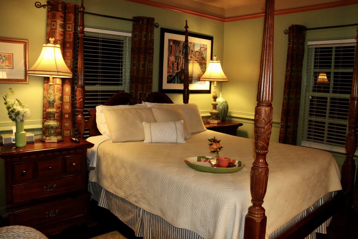 A four-poster bed with a quilted bedspread, a bedside table with a lamp, and a framed picture on the wall.