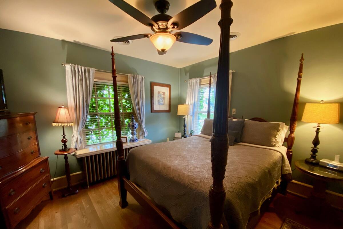 A four-poster bed with a ceiling fan, a dresser, a bedside table with a lamp, and a window with curtains.