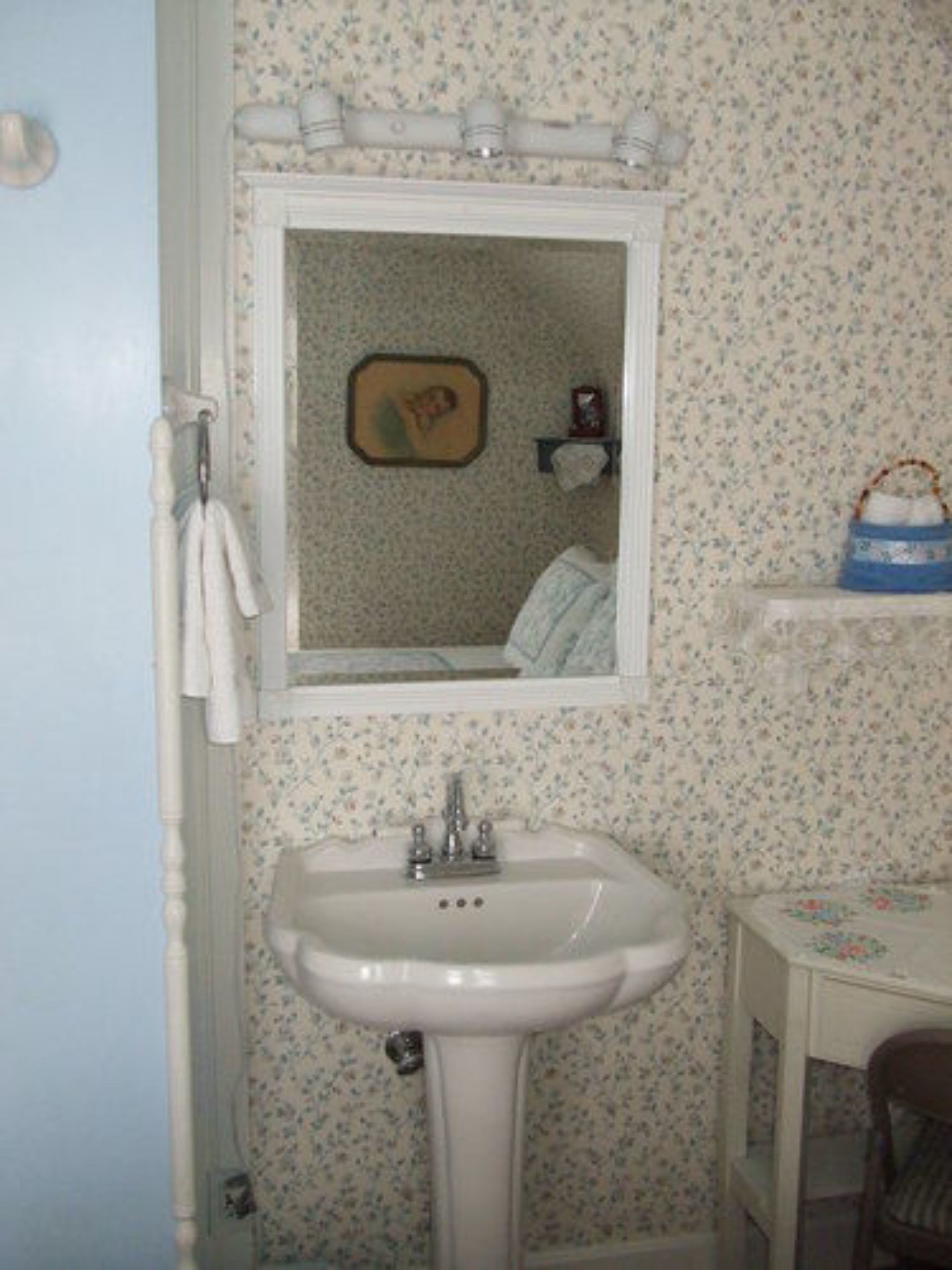 A bathroom with a white sink, a mirror above it, and a floral wallpaper. There is a towel rack on the left and a small table with a basket on the right.