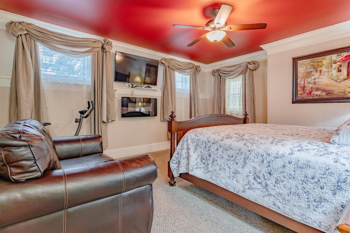 A bedroom with a bed, a chair, a ceiling fan, and a painting on the wall.