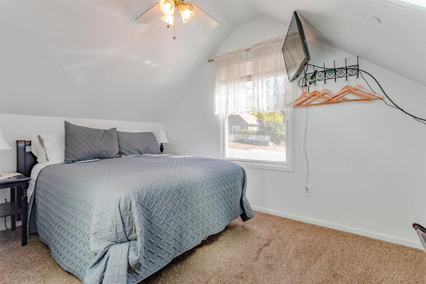 A bedroom with a bed, a nightstand, a lamp, a ceiling fan, a window with sheer curtains, and a wall-mounted TV.