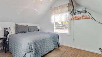 A bedroom with a bed, a nightstand, a lamp, a ceiling fan, a window with sheer curtains, and a wall-mounted TV.