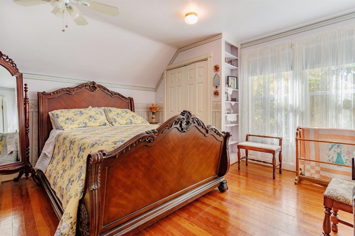 A bedroom with a wooden four-poster bed, a chair, a small table, and a window with sheer curtains.
