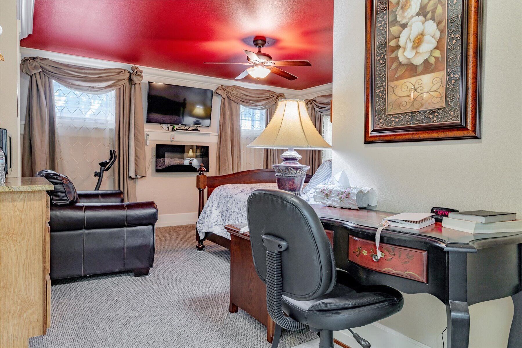 A hotel room with a bed, a desk, a chair, a ceiling fan, a TV, and a framed picture on the wall.