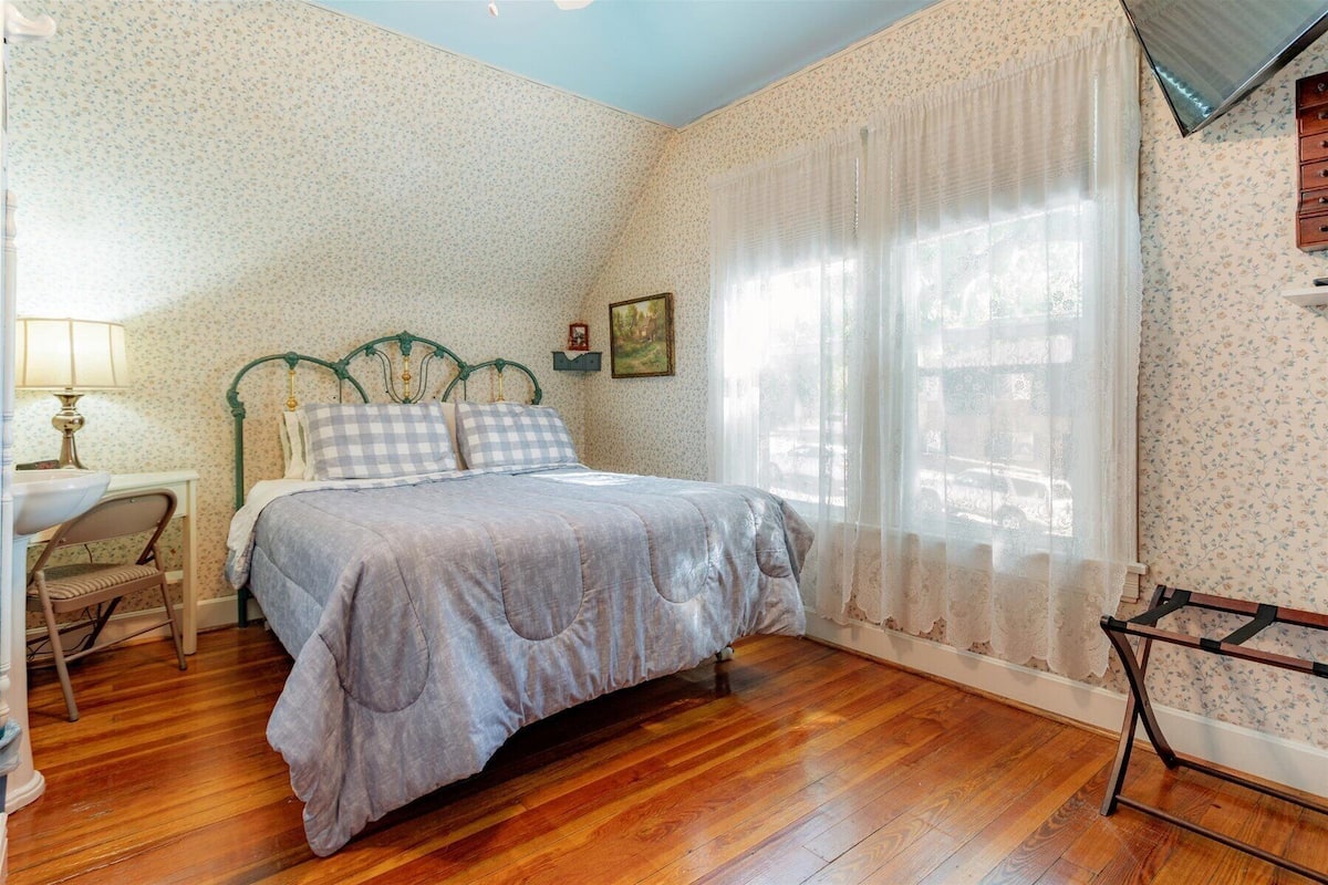 A bedroom with a wooden floor, a bed with a checkered duvet, a nightstand, a lamp, and a chair.