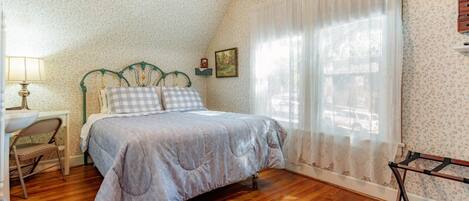 A bedroom with a wooden floor, a bed with a checkered duvet, a nightstand, a lamp, and a chair.