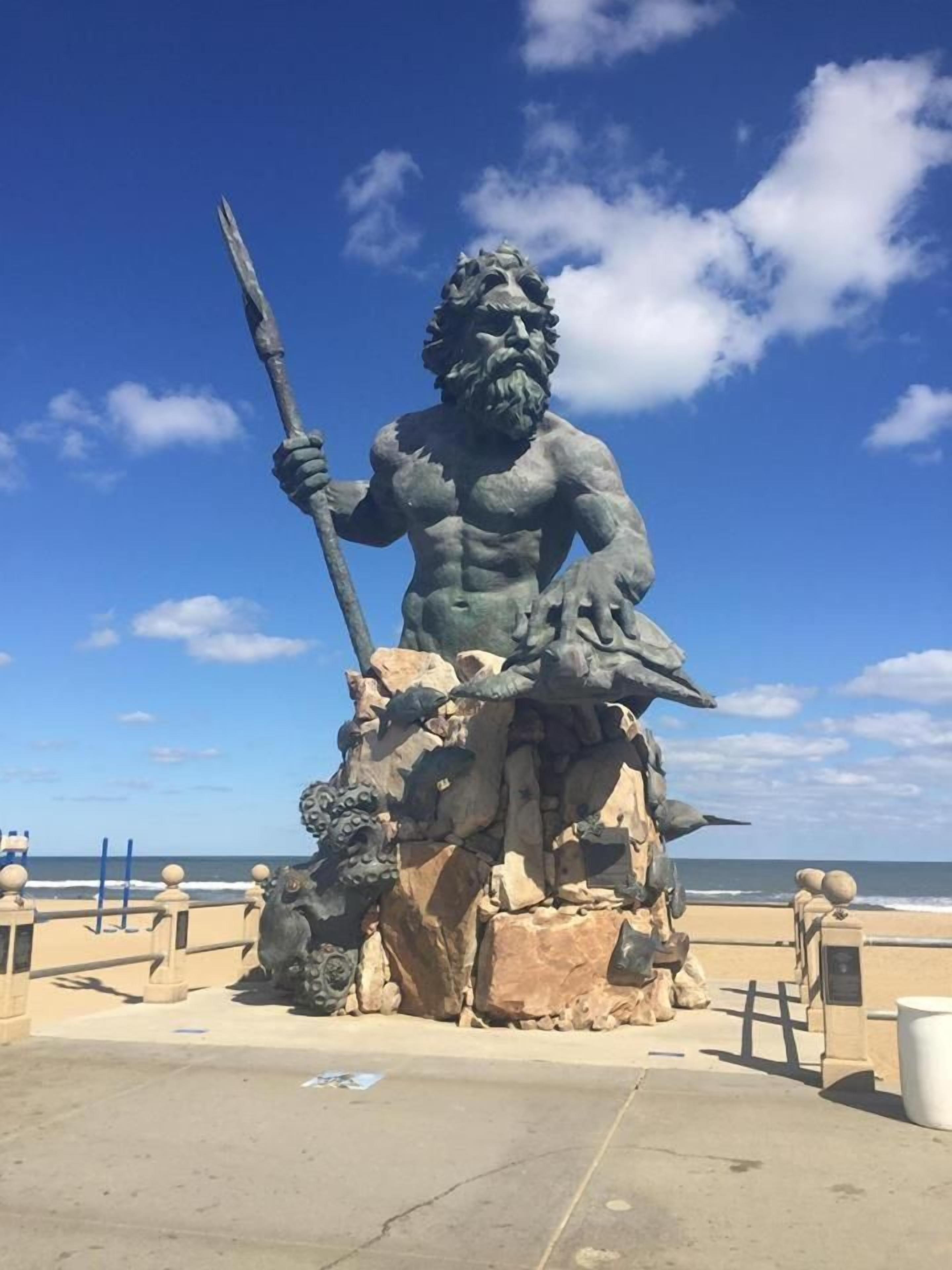 A large bronze statue of a muscular figure with a spear, seated on a rock pedestal, with the ocean in the background.