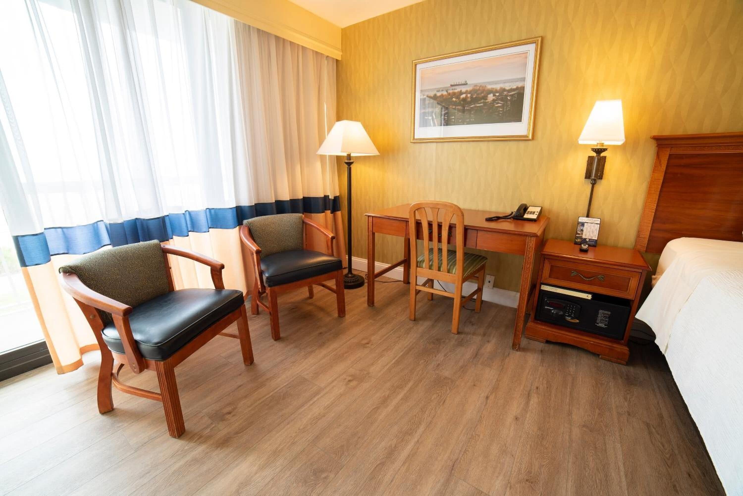 Habitación de hotel con un escritorio de madera, dos sillas, una cama, una mesita de noche con lámpara y un cuadro en la pared.