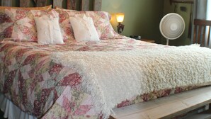 Un dormitorio con cabecera de madera, ropa de cama floral, un candelabro, un ventilador de noche y una ventana con cortina.