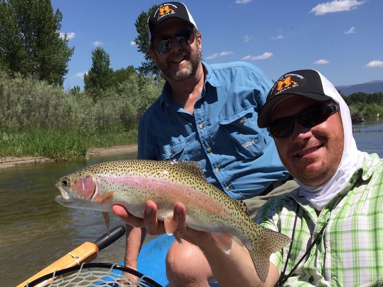 Two people holding a fish, with a river and greenery in the background.
