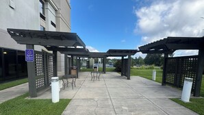 Uma área externa coberta com mesas e cadeiras, sinalização indicando área para fumantes e um edifício ao fundo.
