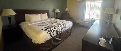 Habitación de hotel con una cama grande, dos mesitas de noche con lámparas, una silla, un escritorio con silla y un ventanal con cortinas.