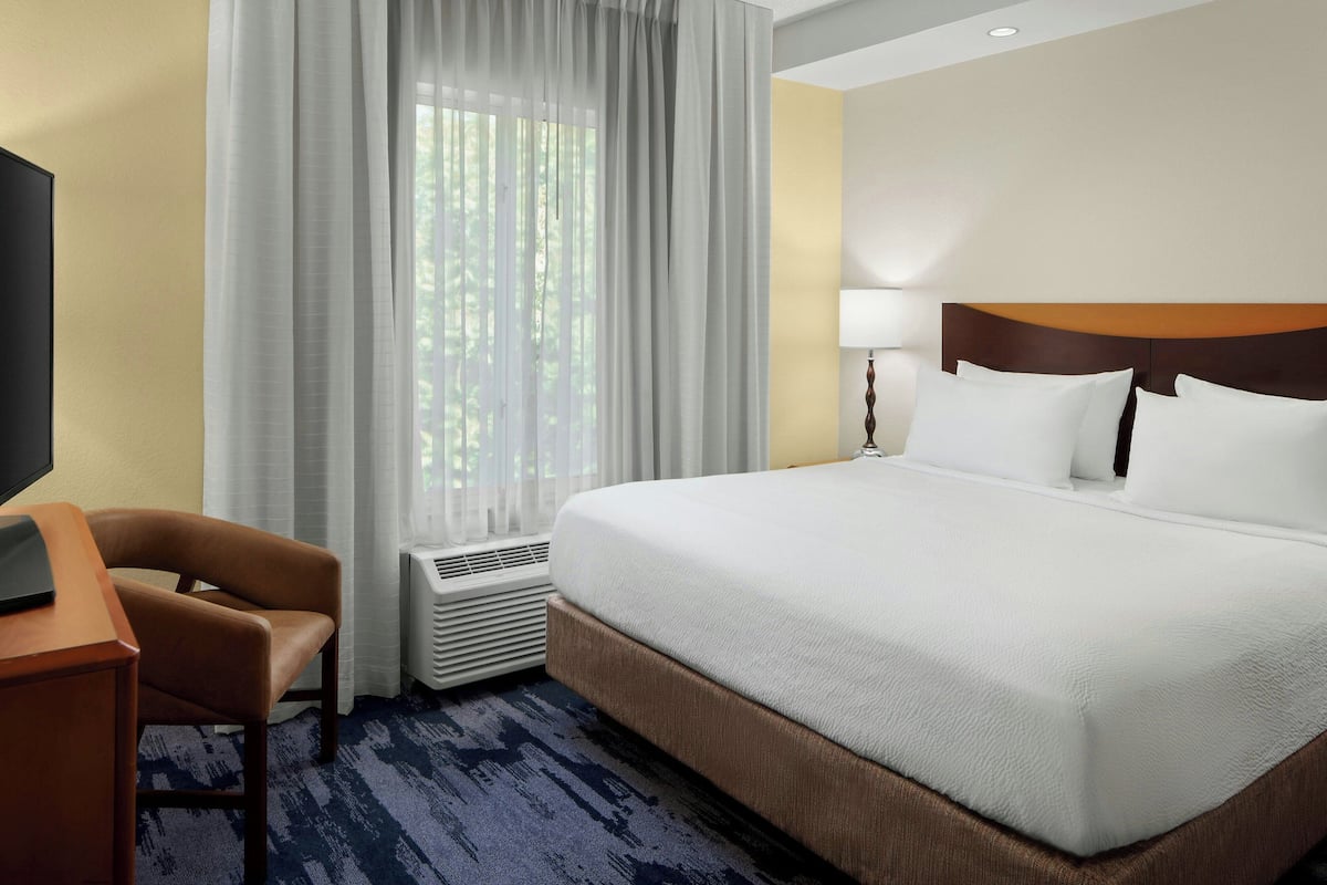 A hotel room with a large bed, a desk, a chair, a window with curtains, and a wall-mounted lamp.