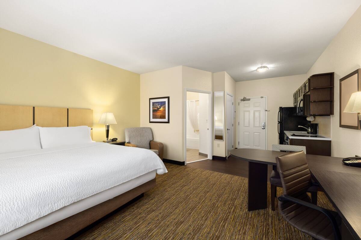 A hotel room with a large bed, a desk, a chair, a kitchenette, and a bathroom.
