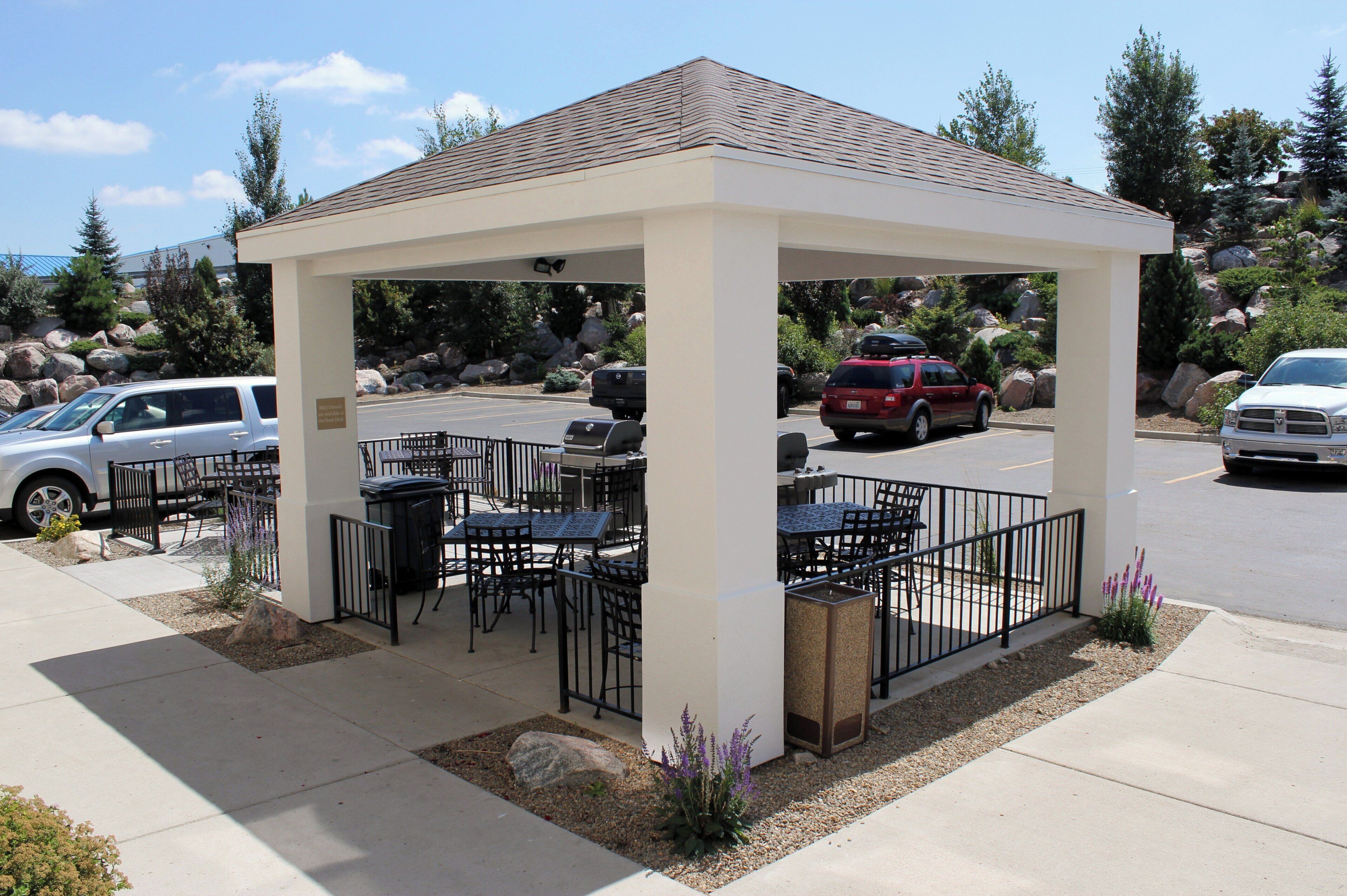 A covered outdoor seating area with tables and chairs, surrounded by a metal railing, set against a backdrop of parked cars and natural scenery.