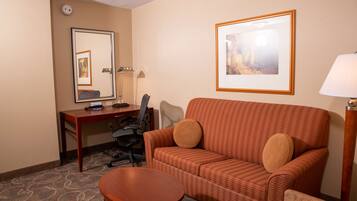 A hotel room with a sofa, a small table, a desk with a chair, a lamp, and framed pictures on the walls.