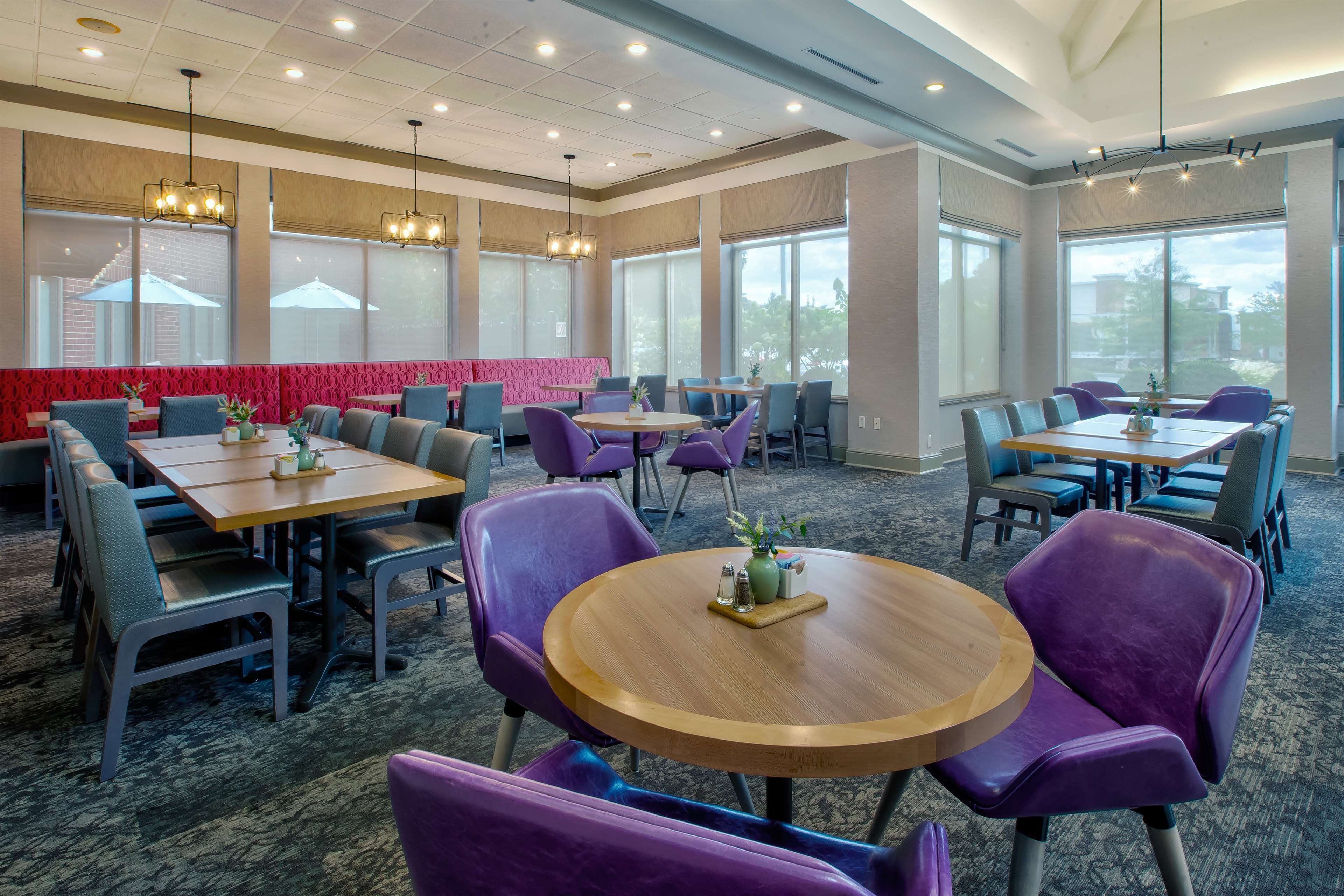 Un espace repas avec des tables rondes et des chaises violettes, de grandes fenêtres et un mur d’accent rouge.