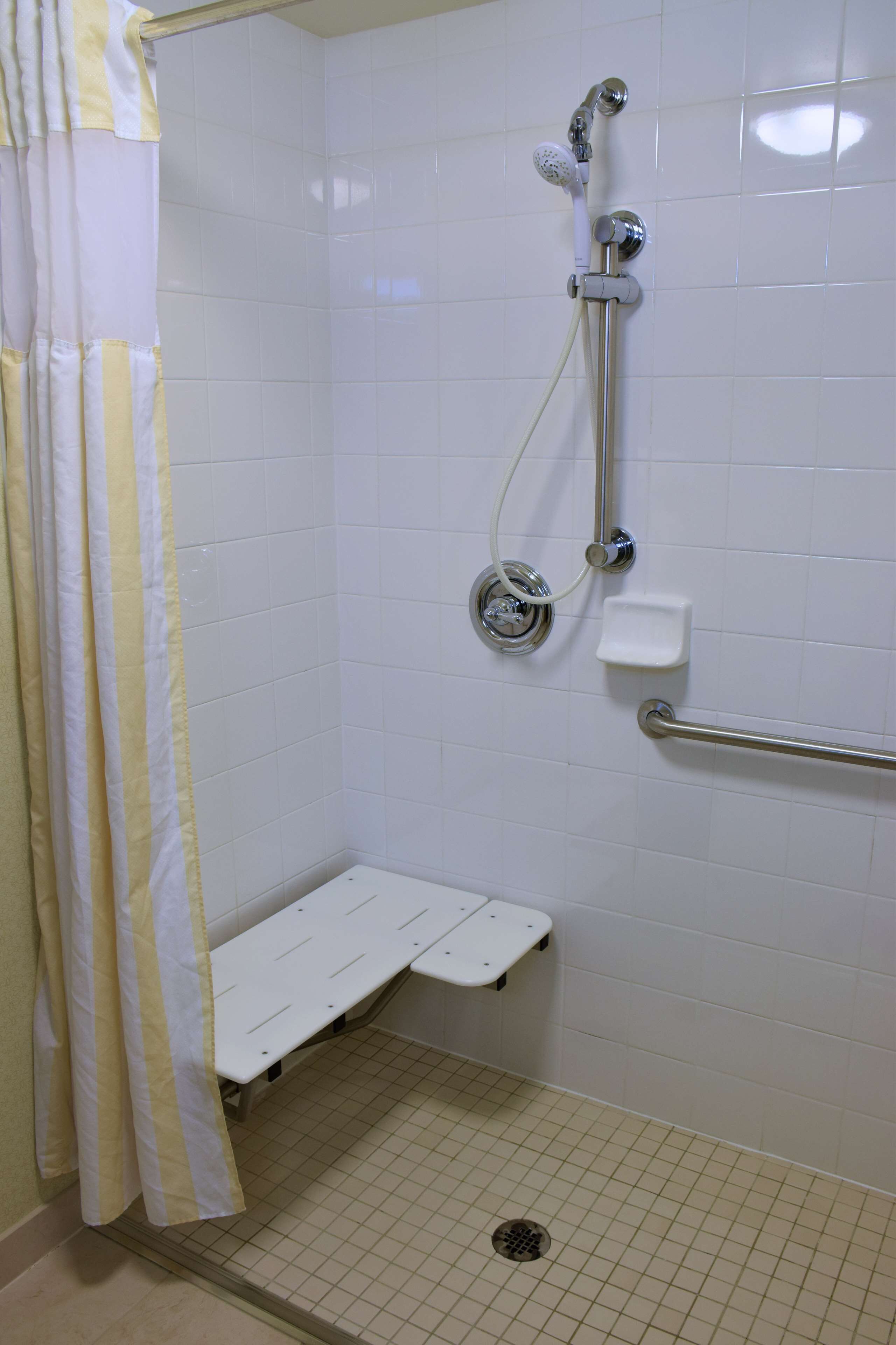 Un espace douche avec un mur recouvert de carreaux blancs, un pommeau de douche, un flexible de douche, un porte-savon et une barre d’appui.