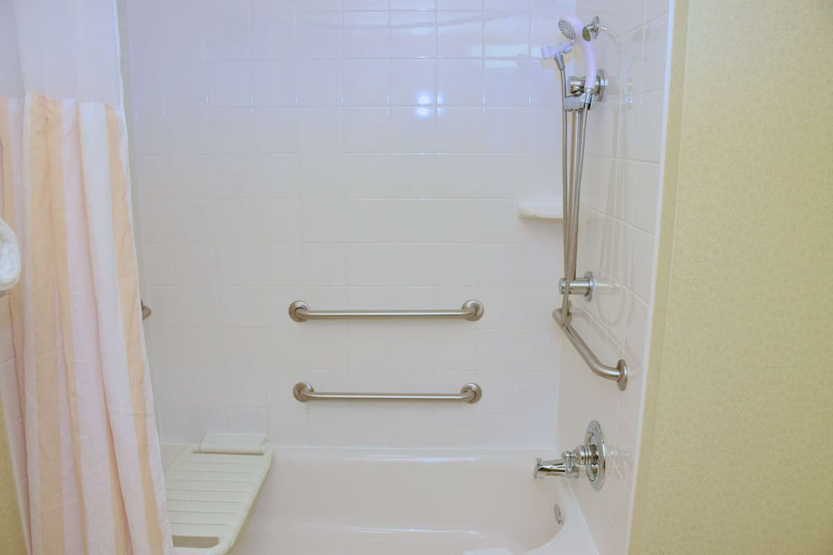 Een badkamer met witte betegelde wanden, een douche met een handdouche en twee handgrepen voor de veiligheid.